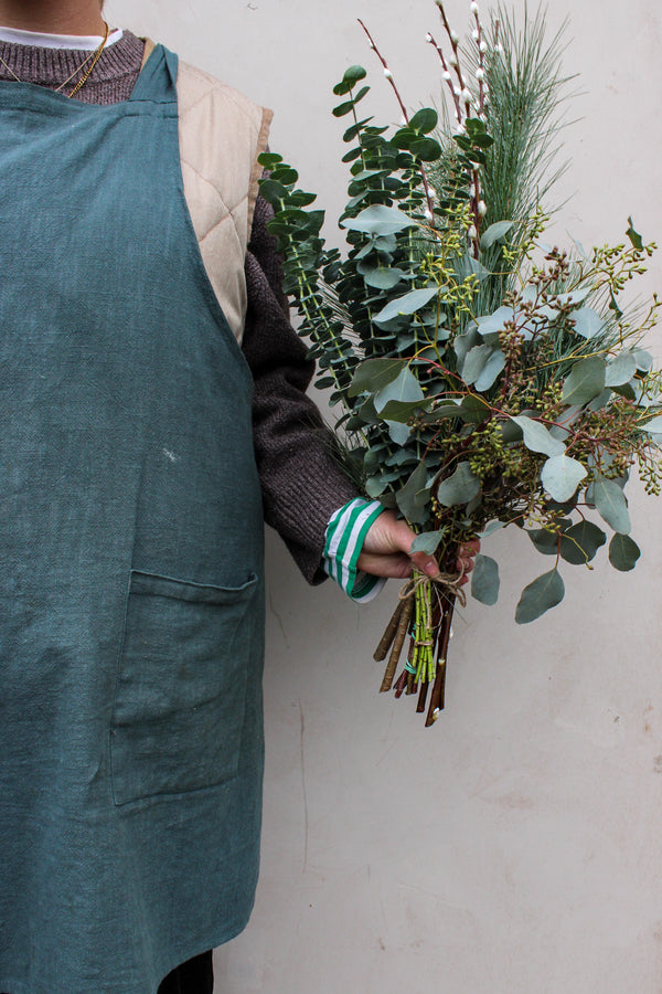 A person in a green apron and brown sweater holds the Wilder One Foliage Stems by Wild About Flowers, featuring festive eucalyptus and pine branches, against a plain light background.