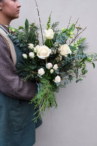 A person in a blue apron and brown sweater holds the Wilder One Christmas Bouquet by Wild About Flowers—featuring white roses, winter flowers, and greenery—set against a light background, perfect for the festive season at Keynsham’s local flower shop.