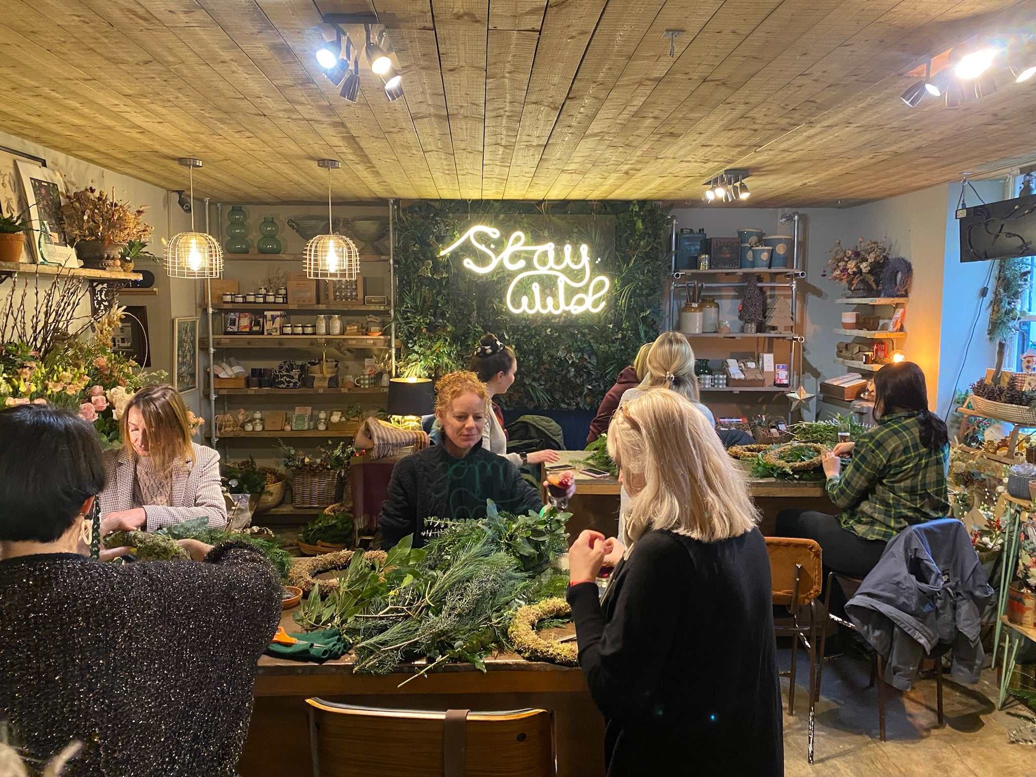 People crafting wreaths at a flower workshop under a "Stay Wild" neon sign